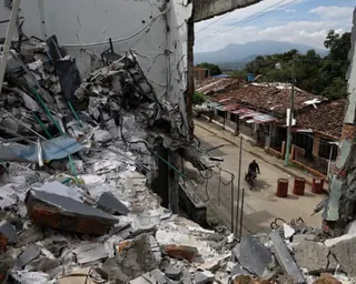 A motorcyclist rides past destroyed buildings with rubble and debris on a street with mountains in the background