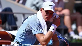 Jannik Sinner, wearing a grey t-shirt and white cap, sits in his chair with a towel around his neck wearing a pained expression
