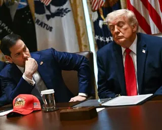 Rubio and Trump, with his eyes closed, sit at a table during a cabinet meeting, with flags and a red cap visible