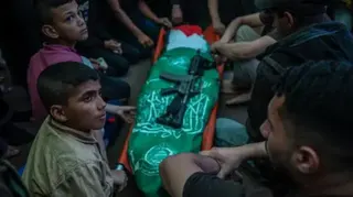 EPA/Shutterstock A body wrapped in flags lying on a stretcher with a gun placed on top of it, and people sitting around the stretcher