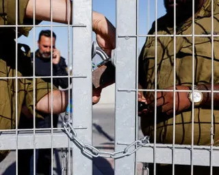 Two soldiers in olive uniforms secure a metal gate with a chain from the inside