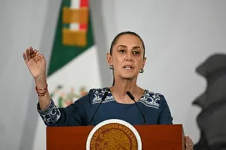 A woman speaks into a microphone with large Mexican flag in the background.