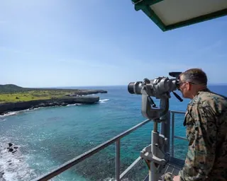 A person in military camouflage looks through mounted binoculars toward a coastal landscape with turquoise water