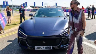 身穿閃亮紫色賽車服的F1賽車手劉易斯漢密爾頓走過停在路邊的一輛法拉利Purosangue SUV。