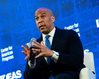 man in suit speaks into microphone while seated on stage