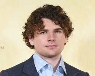 Kane Parsons, a young man with curly brown hair in a dark blazer and light blue shirt, poses for a photo