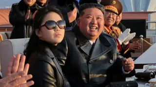 Kim Ju Ae wearing black leather jacket and black sunglasses sitting next to Kim Jong Un who is wearing a black leather trench