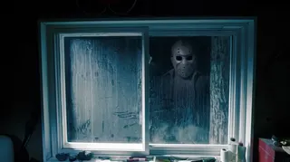 Jason Voorhees wearing a hockey mask is reflected in a grimy window.