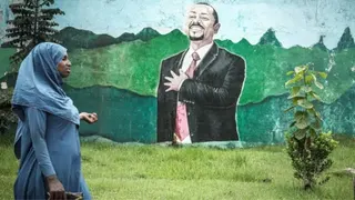 AFP via Getty Images A woman in a blue dress and headscarf walks past grass in front of a wall with a mural of Abiy Ahmed. He is depicted in a black suit, white shirt and red tie holding his arm up over his heart.