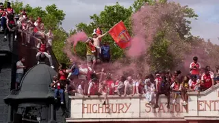 PA Media A large group of Arsenal football fans celebrate whilst gathered on the roof and structure of Highbury Station. Several supporters wear the club's red and white shirts, one fan waves a large Arsenal flag, and another holds a flare that releases thick plumes of pinkish-red smoke into the air.