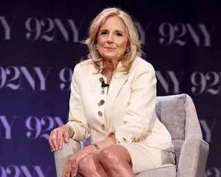 Jill Biden in a cream suit with gold buttons sits in a chair and smiles at the camera