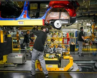 Workers assemble a suspended car chassis with red bodywork on a factory production line