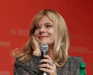 Nastassja Kinski holds a microphone and rests her chin on her hand, smiling warmly