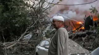 EPA A man wearing a grey top stands with his head bandaged as he surveys the aftermath of an attack. Beside him is a silver car, whose roof is covered with debris, trees that have fallen down in what appears to be a forested area and where a fire is still burning bright