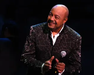 A man in a black and gold velvet jacket on stage, smiling and clapping.