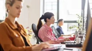 Getty Images A row of workers looking at their laptops in a brightly lit office. The icture focuses on a woman looking at a monitor in a red and white striped blouse, with her dark hair in a ponytail.