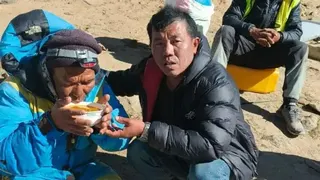 一名身穿藍色登山外套的男子正在用碗喝橙色湯品，旁邊有一名穿黑色羽絨外套的男子在協助他