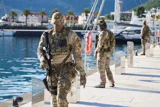 Armed personnel in camouflage, carrying guns, patrol on a harbourfront. There are yachts in the background.