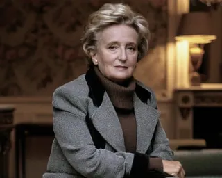Bernadette Chirac in a grey jacket with black collar and brown turtleneck sits in an elegant interior