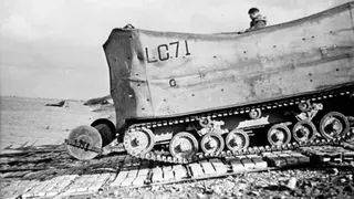 A black and white photo shows a profile view of a large World War Two tank on caterpillar tracks, descending into a river on a rolled-out ramp. A soldier can be seen looking over the top, towards the water.