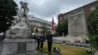 Royal British Legion personnel salute the memorial which has several wreaths laid at the bottom.  The memorial is a large oblong stone statue standing upright.A large stone statue of two army personnel is in the foreground.