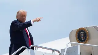 US President Donald Trump, at the top of a staircase up to Air Force One, pointing to the right. He is wearing a navy blue suit, white short and red neck tie.