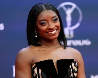 Simone Biles in a black dress smiles at the camera in front of a blue backdrop