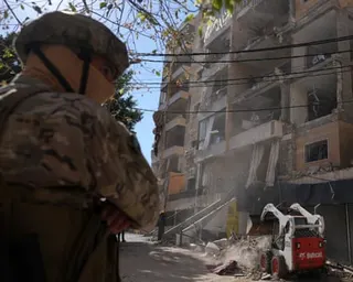 A soldier in full combat gear stands next to a badly damaged apartment block