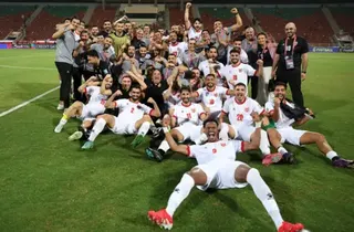 Ameen Ahmed/NurPhoto via Getty Images A group picture of Jordanian football players and other supporting members of the wider team smiling and celebrating on a football pitch.  Many of the players are wearing white kit and sitting on the ground. They are all looking at the camera.