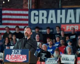 Graham Platner speaks at a podium with a 'Fight Oligarchy' sign in front of a crowd