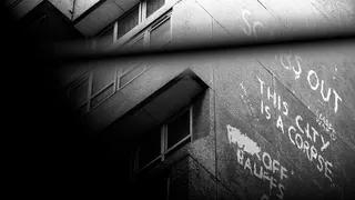 A black and white image showing the side of a block of flats with graffiti on. The words "this city is a corpse" can be clearly seen.