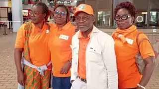 Julien Kouadio Adonis A man wearing an orange shirt and baseball cap and a white jacket looks at the camera. He has glasses and short facial hair. Three women stand with him, also wearing orange polo shits.