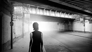 An artfully framed screenshot of the underside of a bridge in a city, but there's only one character walking down the street and it's all coloured in black and white.