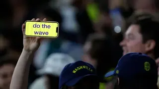Getty Images A person whose face we cannot see wears a blue baseball cap and holds aloft a mobile phone with the words 'do the wave' on it. There is a blurry photo in the background of someone smiling. This appears to be a sports stadium full of people.