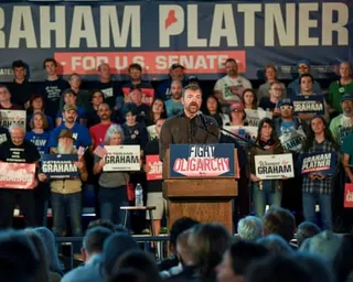 man speaks at lectern marked 'fight oligarchy' as crowd holds signs saying 'women for graham', 'graham platner' and 'veterans for graham'