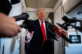 man in suit speaks into reporters' microphones while on plane