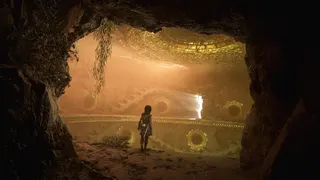 A curly haired woman stands in a darkened interior looking at a chamber filled with eye-like symbols. It's Sophia in Resonance: A Plague Tale Legacy.