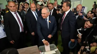 Reuters A man puts his ballot in a box while smiling at people off to the side. He is surrounded by men in suits and members of the press