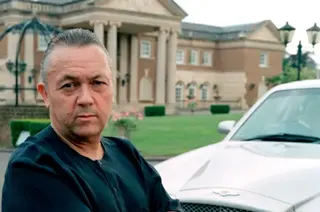 David Sullivan, wearing a black T-shirt, poses with his arms folded next to a silver Bentley outside his home, Birch Hall in Essex, a large neo-classical mansion with an imposing stone portico supported by pillars. He was pictured in 2003 for a BBC documentary called Sex Empires: Rise of the Porn Barons