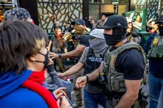 a man in tactical gear holds a taser as other men in tactical gear stand in a crowd of people on a street