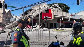 Policemen in uniform stand in front of a barricaded road. Behind them is a partially collapsed building with a red Jollibee sign.