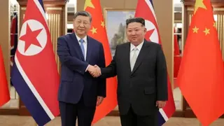 Xi Jinping and Kim Jong Un shaking hands. Both are wearing suits. Behind them is a row of Chinese and North Korean flags.