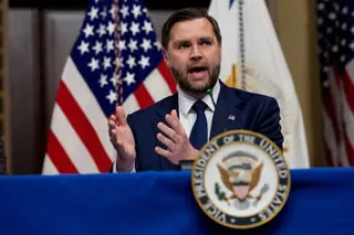 A man speaks into a microphone while seated at a table with a US flag as a backdrop.