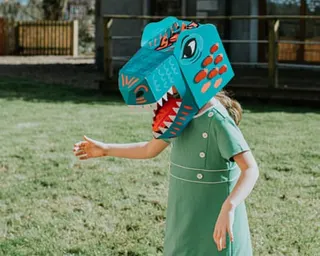 A girl in a domestic garden wears a brightly coloured cardboard dinosaur mask