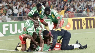 Cameroon outfield players in green shirts, red shorts and yellow socks, as well as the goalkeeper dressed mostly in black including his tracksuit bottoms, celebrate a goal at the 1990 World Cup by piling on top of Eugene Ekeke, who lies flat on his back as six players gather around him and offer congratulations. Advertising hoardings can be seen in the background, with a packed stand of fans out of focus in the distances. One player, Roger Milla, looks up from the pack and smiles towards the cam