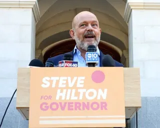 A man speaking to peopl from behind a lectern. A yellow board in front reads 'Steve Hilton For Governor'.