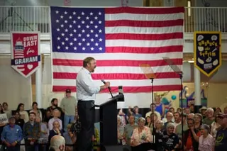 Side-on view of Platner on a podium speaking to crowds, in front of a US flag