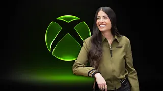 A woman in a thin green jacket and with long black hair an a very white smile leans against an Xbox Logo while smiling widely. It's Xbox boss Asha Sharma.