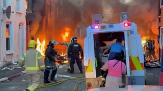 PA Media A riot-control police vehicle is parked in a narrow street as firefighters battle multiple fires around burning cars and buildings. Thick smoke fills the area while emergency crews and a few civilians move through the scene amid debris and fire hoses.