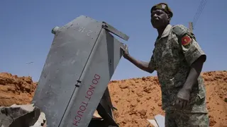 Los Angeles Times via Getty Images A man in a military uniform stands in a crater, surrounded by sand. His right hand rests on the fin of a downed drone which says "do not touch".